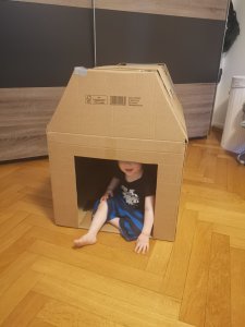 Haus aus Pappe. Kleinkind sitzt lächelnd drinnen.