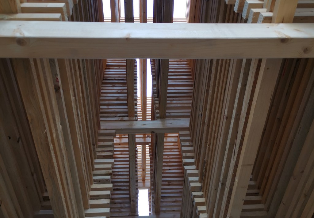 Holzbalken, die zu einem Turm geformt sind. Blick von Innen hinauf in den Himmel.